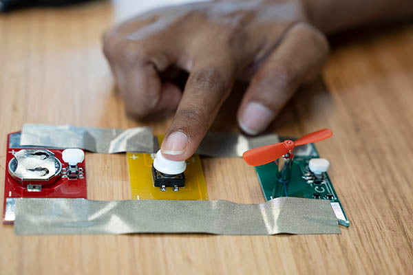 A red, yellow and green circuit board they are taped to a table with grey tape. The red board has a battery, the middle board has a white button and the green one has a fan.