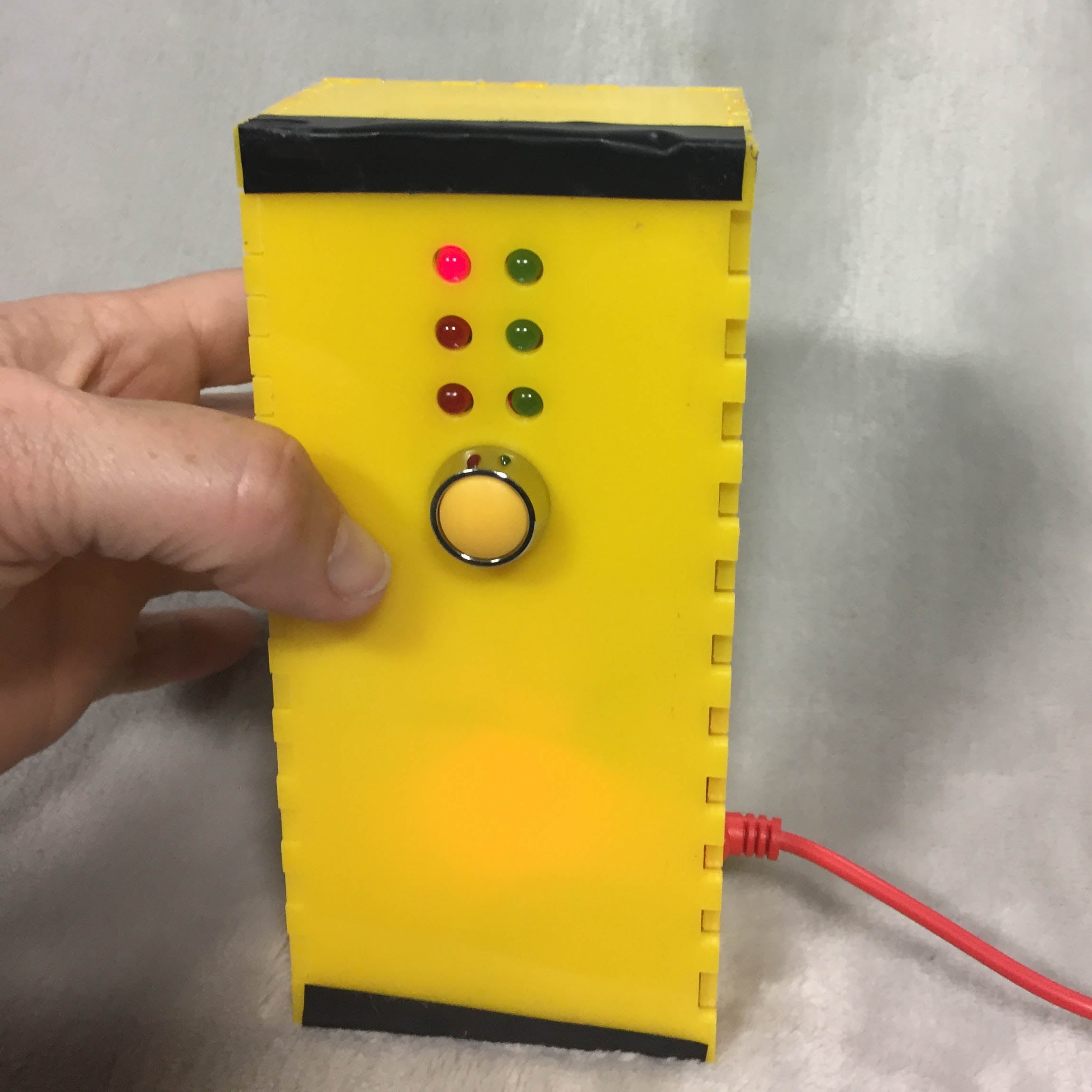 A yellow box with three red and three green light arranged as a braille cell. A yellow buttons is in the middle