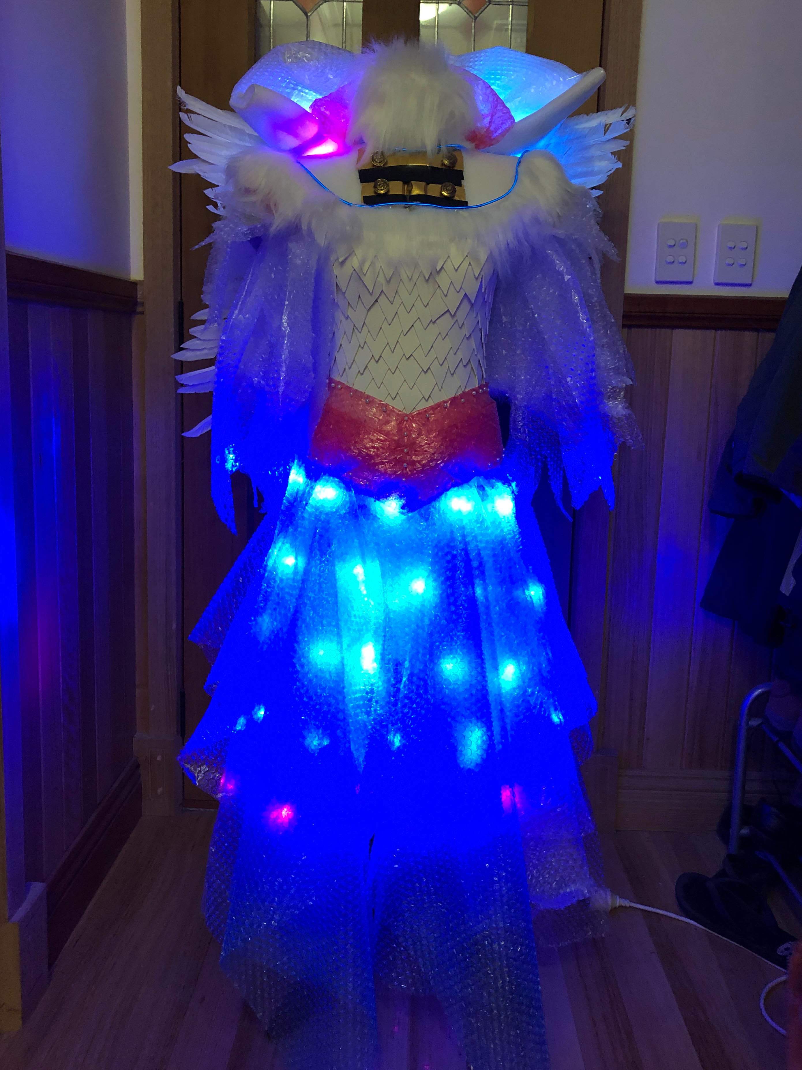 A model in a white dress that is made of bubble wrap with a foam tile bodice.  The dress is lit up by blue LED lights in the collar, arms and skirt. There is a red belt and blue front decorative section.