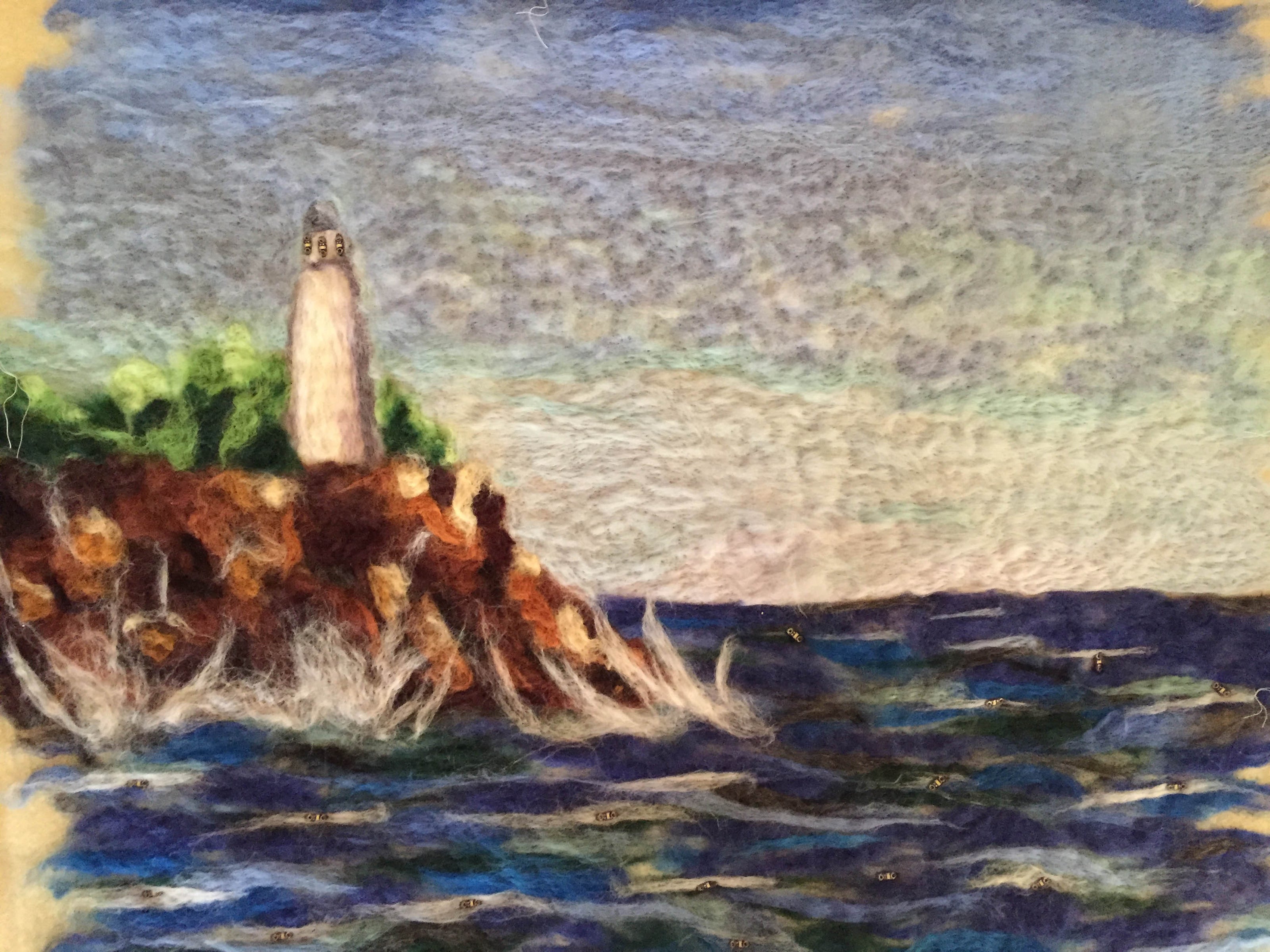 A dry felted artwork with a lighthouse on the left on top of cliffs and rough blue water at the front.