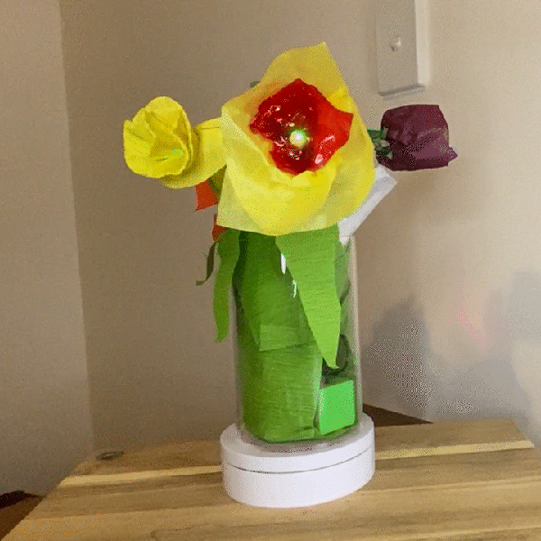 A vase with 4 paper flowers on a rotating stand. The centre flower is red and yellpw and has a green centre light