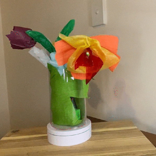 A vase with 4 paper flowers on a rotating stand. The centre flower is red, yellow and  orange and has a light in the centre
