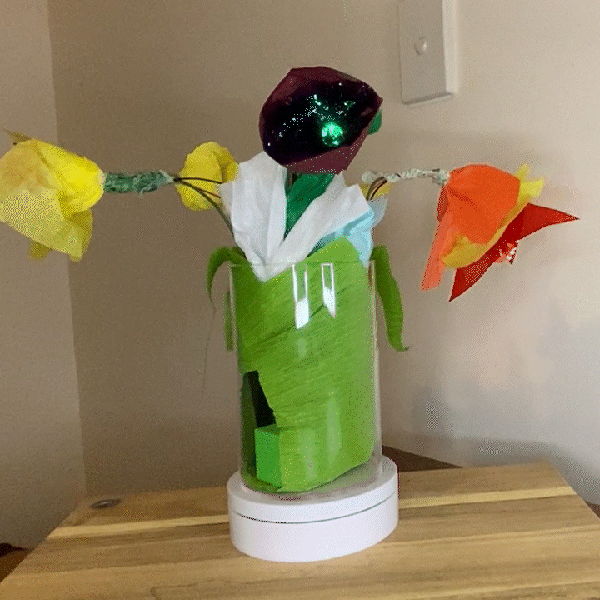 A vase with 4 paper flowers on a rotating stand. The centre flower is purple and has a green centre light