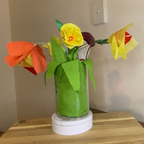 A vase with 4 paper flowers on a rotating stand. The centre flower is yellow and has a red centre light