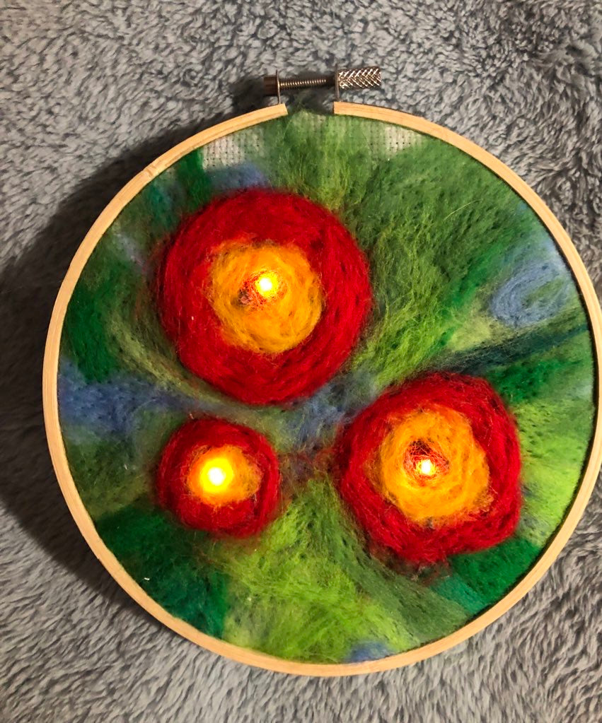 A round dry felted picture with three red and yellow flowers with yellow lights in the middle