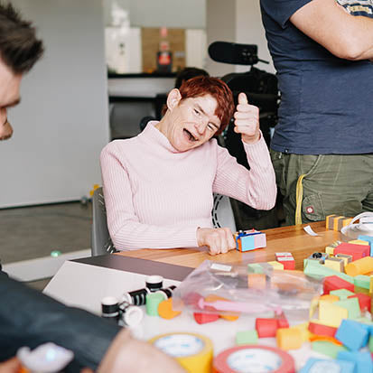 A phone of a lady with a disability with her thumb up smiling with a TapeBlock circuit in front of her that she has made.