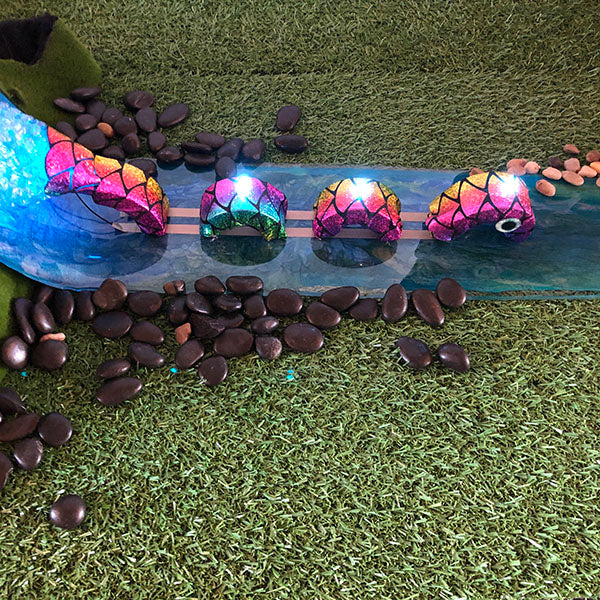 A colorful material serpent with lights on its coils appears to be swimming along a river which is made of resin. It is surrounded by rocks and grass