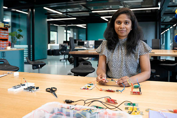 A brown woman sitting at a wooden table holding a circuit and has several other boards around. In the background is a green open office