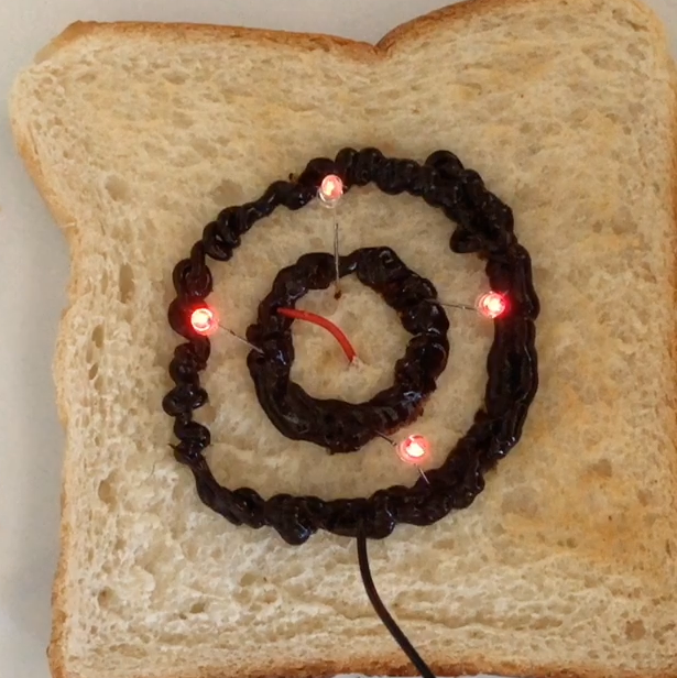 A piece of toast with two rings of Vegemite, one inside the other (not touching) there are four red LEDs that are connected to both rings of Vegemite. A red wire and a black wire are connected to the Vegemite rings