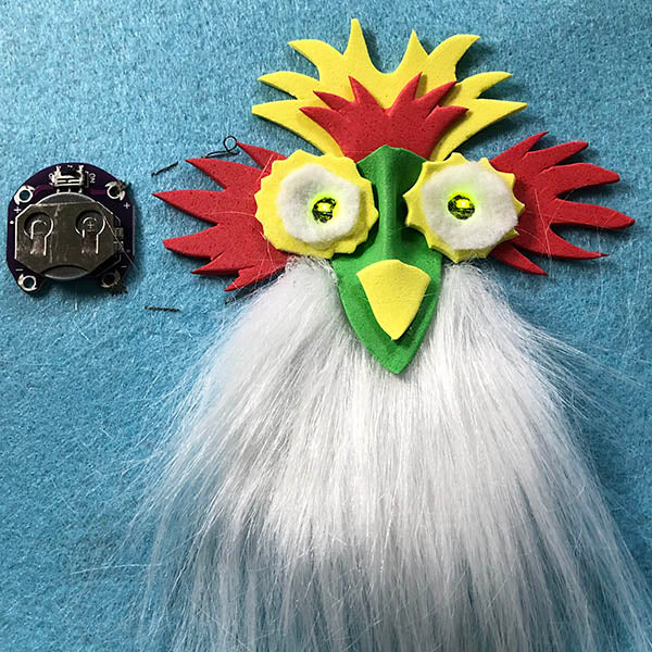 On the left is a circular lily pad button cell battery carrier. This is connected with grey thread  top and bottom to two yellow-yellow lily pad LED which form the eyes of a chook looking creature that has red and yellow decorations and a white beard.